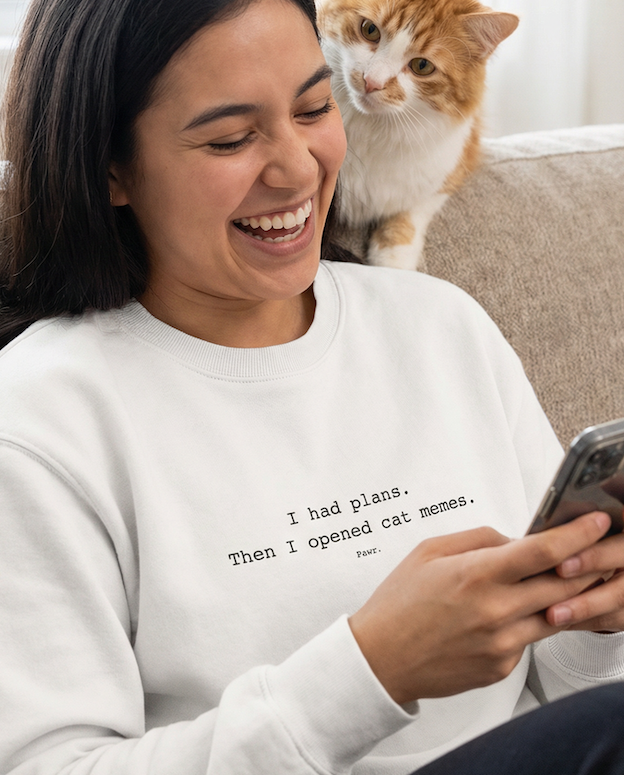 Woman sitting on a couch with a cat on her lap, using a phone, wearing a sweatshirt with text.