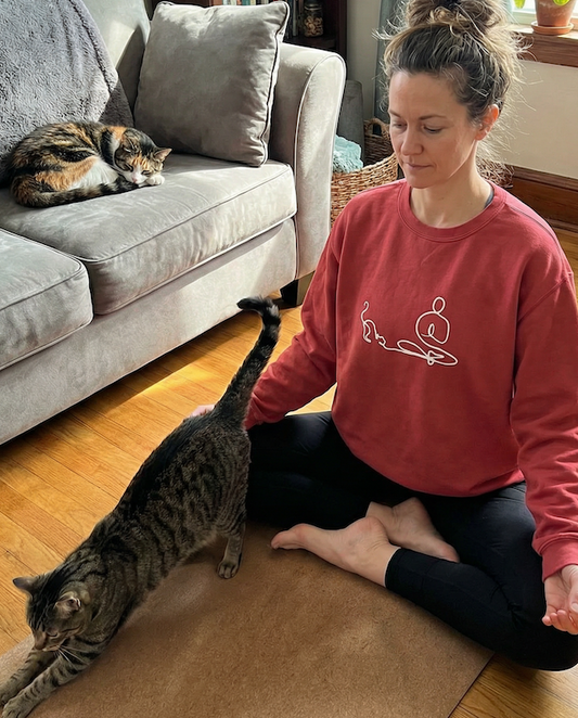 Woman in a red sweatshirt meditating on the floor with a cats in a living room.