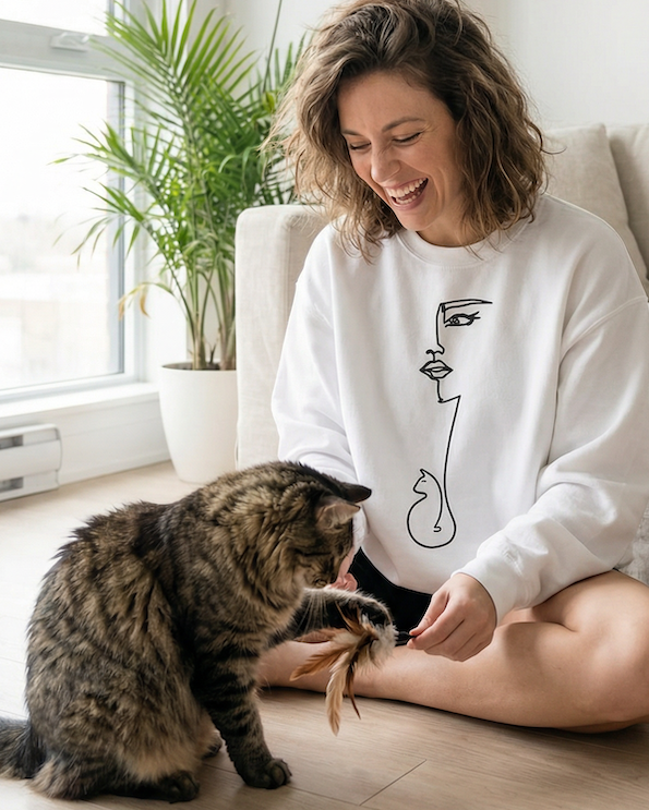 Woman playing with a cat on a wooden floor in a bright room with plants and a couch.