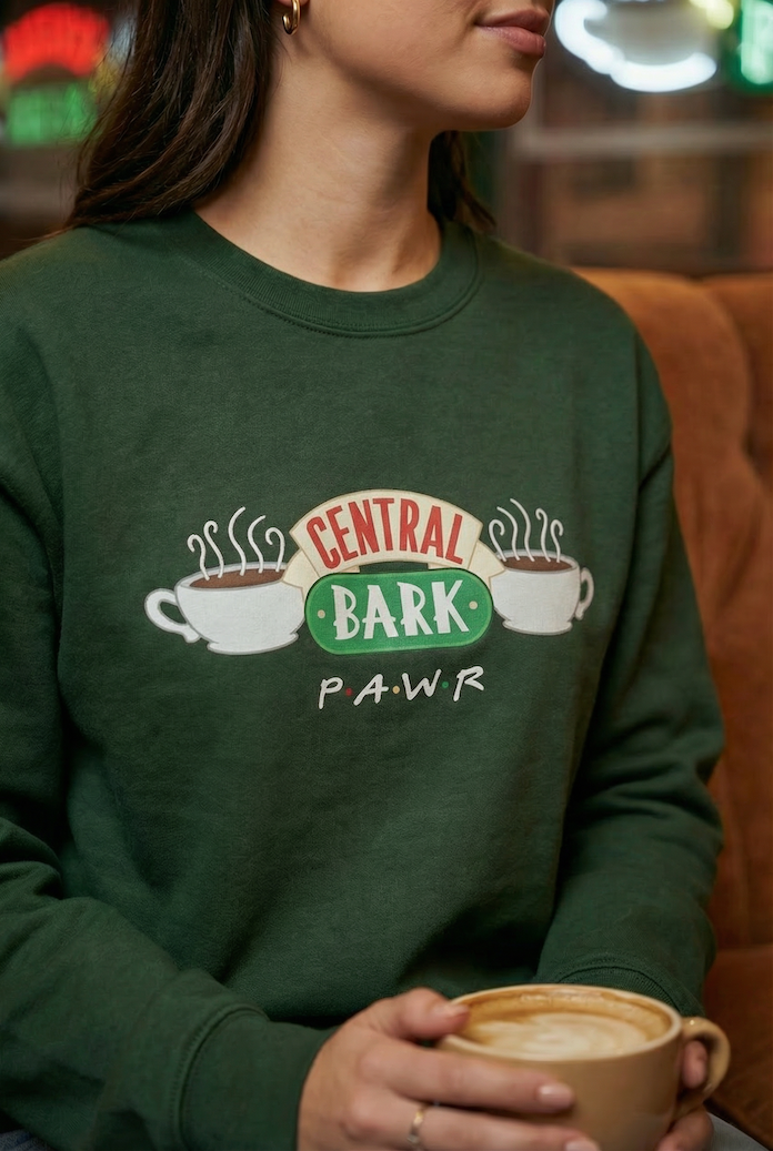 Person wearing a green sweatshirt with 'Central Bark PAWR design, holding a coffee cup.