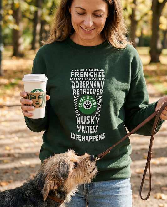 Woman in a green sweatshirt with dog names holding a coffee cup and walking a dog in a park.