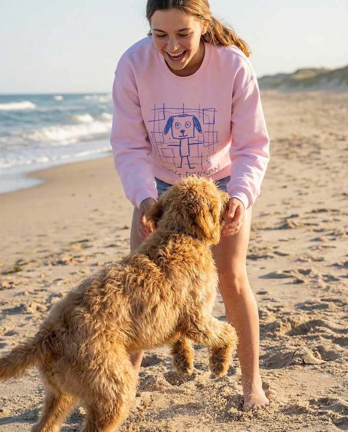 Woman in pink sweatshirt playing with a dog on a sandy beach.