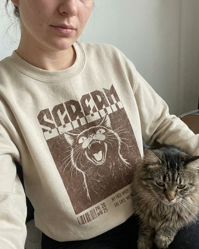 Woman wearing a PAWR sarcasm cat sweatshirt with a vintage-style screaming cat graphic, sitting with a tabby cat on her lap, perfect for cat moms, cat lovers, and cozy everyday wear.