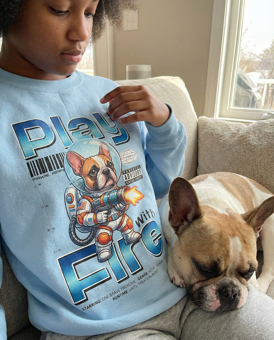Person wearing a light blue PAWR French bulldog sweatshirt with an astronaut Frenchie graphic, sitting on a couch with a real French bulldog, cozy home setting for Frenchie lovers.