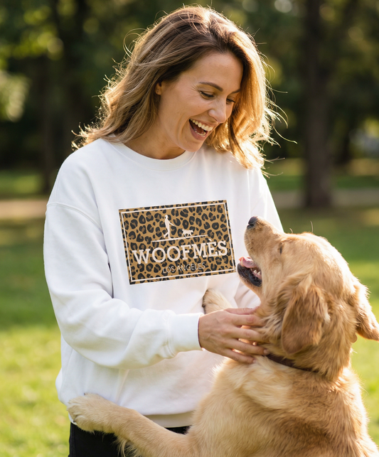 Woman smiling outdoors wearing a white PAWR Woofmes dog sweatshirt with a leopard-style graphic, playing with her dog in a park setting, cozy unisex fit for dog moms and dog lovers.