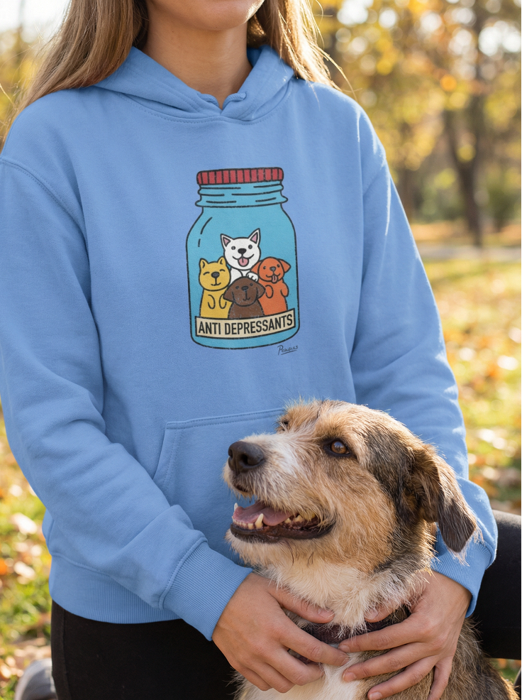 Light blue dog hoodie with “Anti-Depressants” jar graphic featuring four cartoon dogs, worn by a woman kneeling outdoors with her smiling dog in autumn park setting.