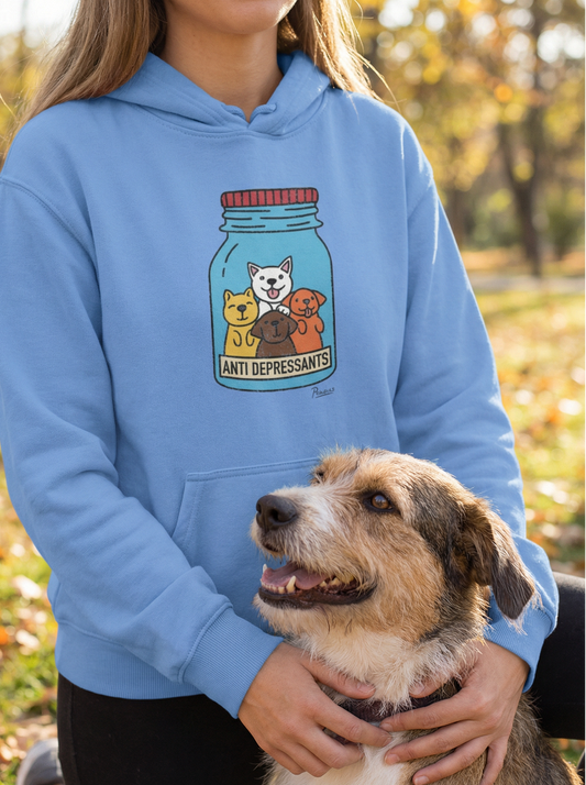 Light blue dog hoodie with “Anti-Depressants” jar graphic featuring four cartoon dogs, worn by a woman kneeling outdoors with her smiling dog in autumn park setting.