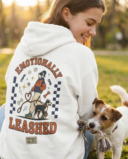 Person wearing a white PAWR Emotionally Leashed dog hoodie with a dog-walking illustration on the back, smiling outdoors while playing with a dog in a park setting.