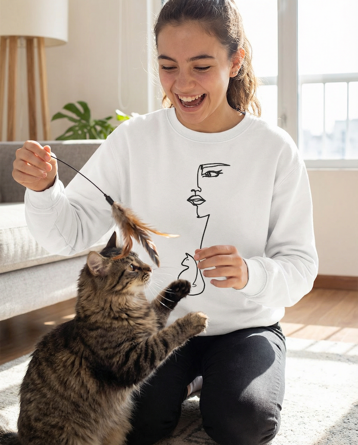 Woman playing with her cat indoors wearing a white PAWR minimalist cat lover sweatshirt with line art face and cat illustration, cozy unisex fit, perfect for cat moms and cat lovers.