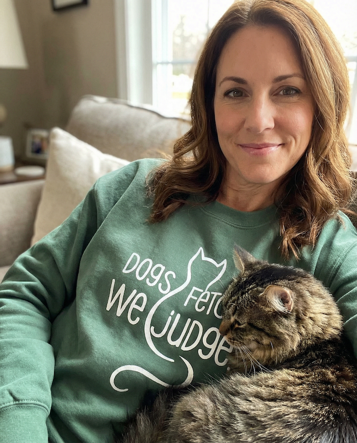 Woman wearing a green sweatshirt with a cat-themed design, sitting with a cat on her lap in a living room.
