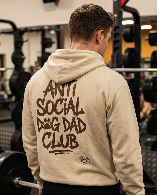 Man wearing a beige PAWR Anti Social Dog Dad Club hoodie with bold back text graphic in a gym, showcasing streetwear-style dog dad apparel designed for dog lovers, workouts, and everyday wear.