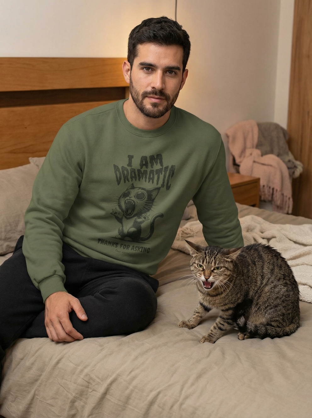 Man wearing green I am dramatic cat sweatshirt sitting on bed with angry tabby cat