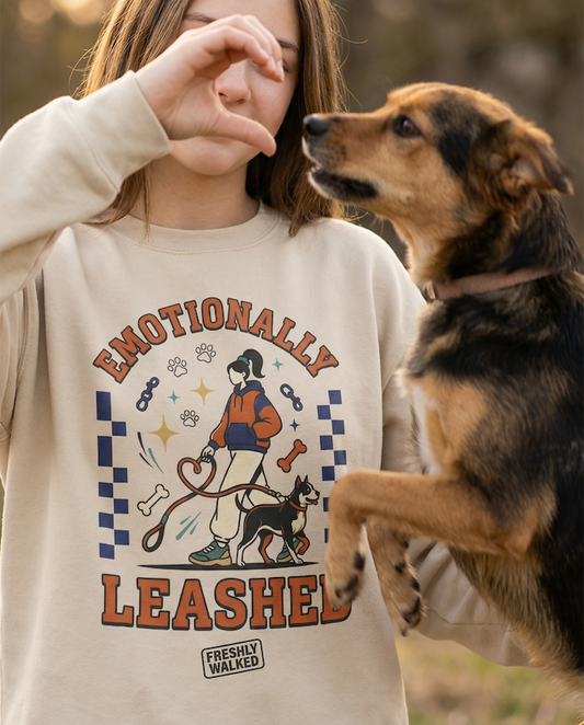 Person wearing a PAWR Emotionally Leashed dog sweatshirt with a playful dog-walking illustration, holding a heart gesture while a dog stands beside them outdoors, perfect for dog lovers.