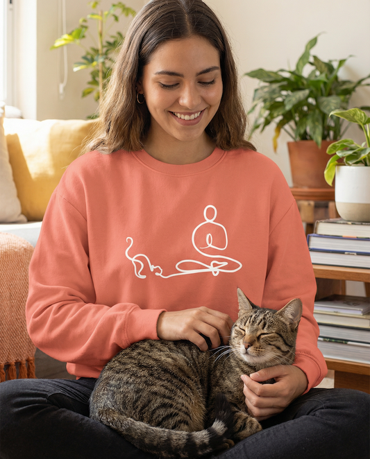 Woman sitting indoors wearing a coral PAWR minimalist cat lover sweatshirt with line art cat design, petting a tabby cat on her lap, cozy unisex fit for cat moms and cat lovers.