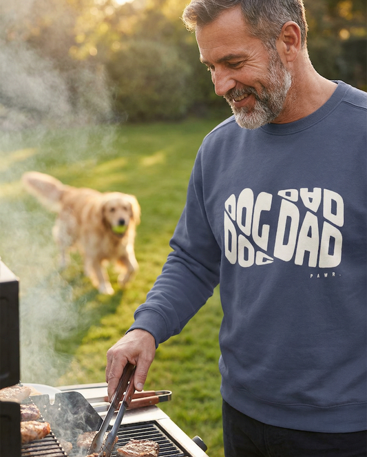 Man grilling outdoors wearing a blue PAWR Dog Dad sweatshirt while his dog plays in the yard, cozy unisex fit, bold Dog Dad text, perfect for proud dog dads and dog lovers.
